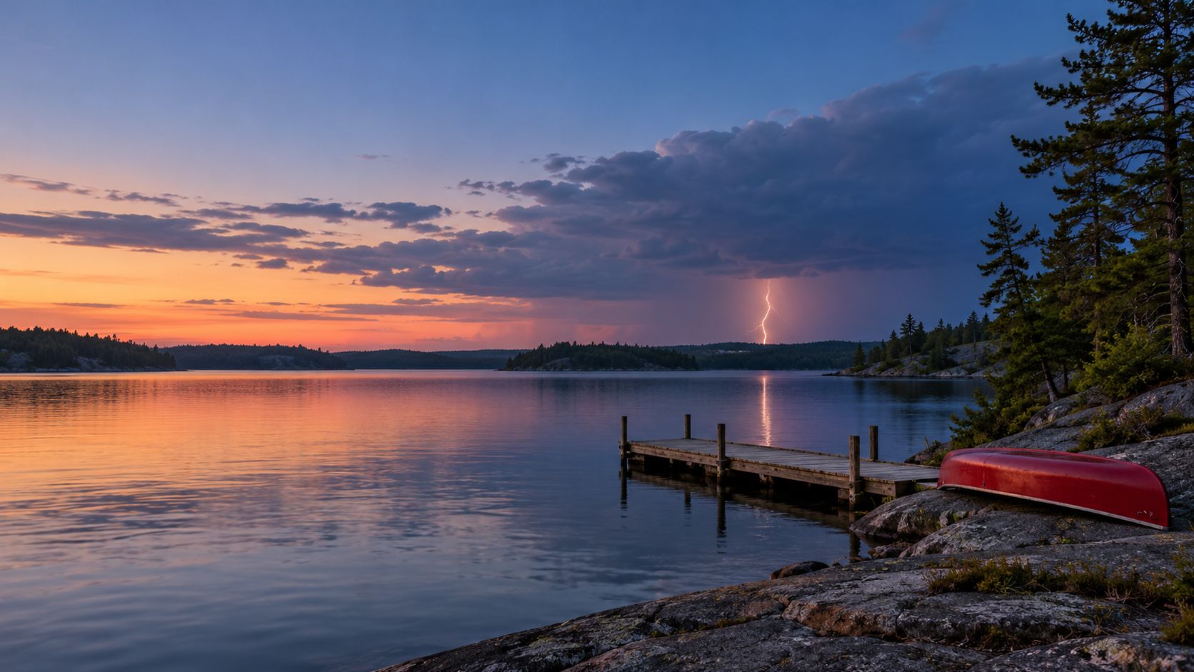 A wooden dock and an overturned red canoe on the rocky shore of a calm Canadian lake at dusk, with lightning from a distant storm over the far shore